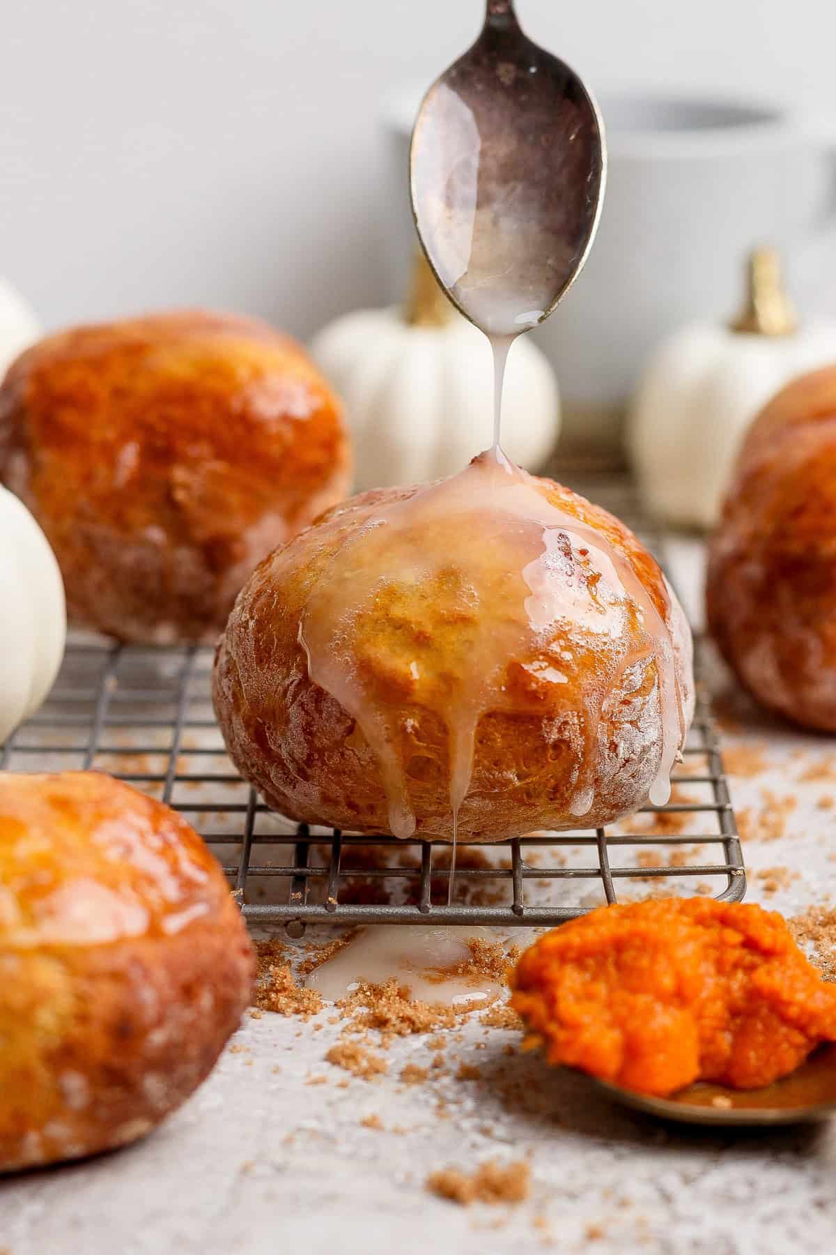 Spoon drizzling white glaze over a round pumpkin fritter on a cooling rack with more fritters and pumpkins behind it.