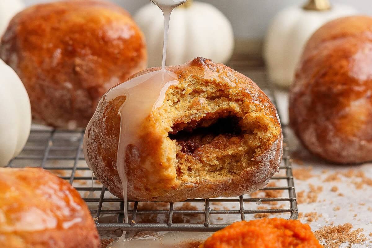 Glazed cottage cheese pumpkin fritter with a bite removed to show the spiced brown sugar center, surrounded by more fritters and small pumpkins.