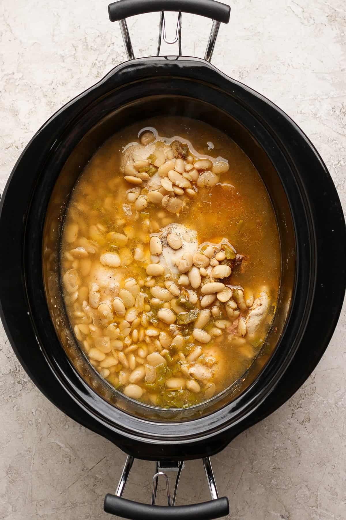 Cooked chicken and white beans in their cooking liquid inside a slow cooker before shredding.