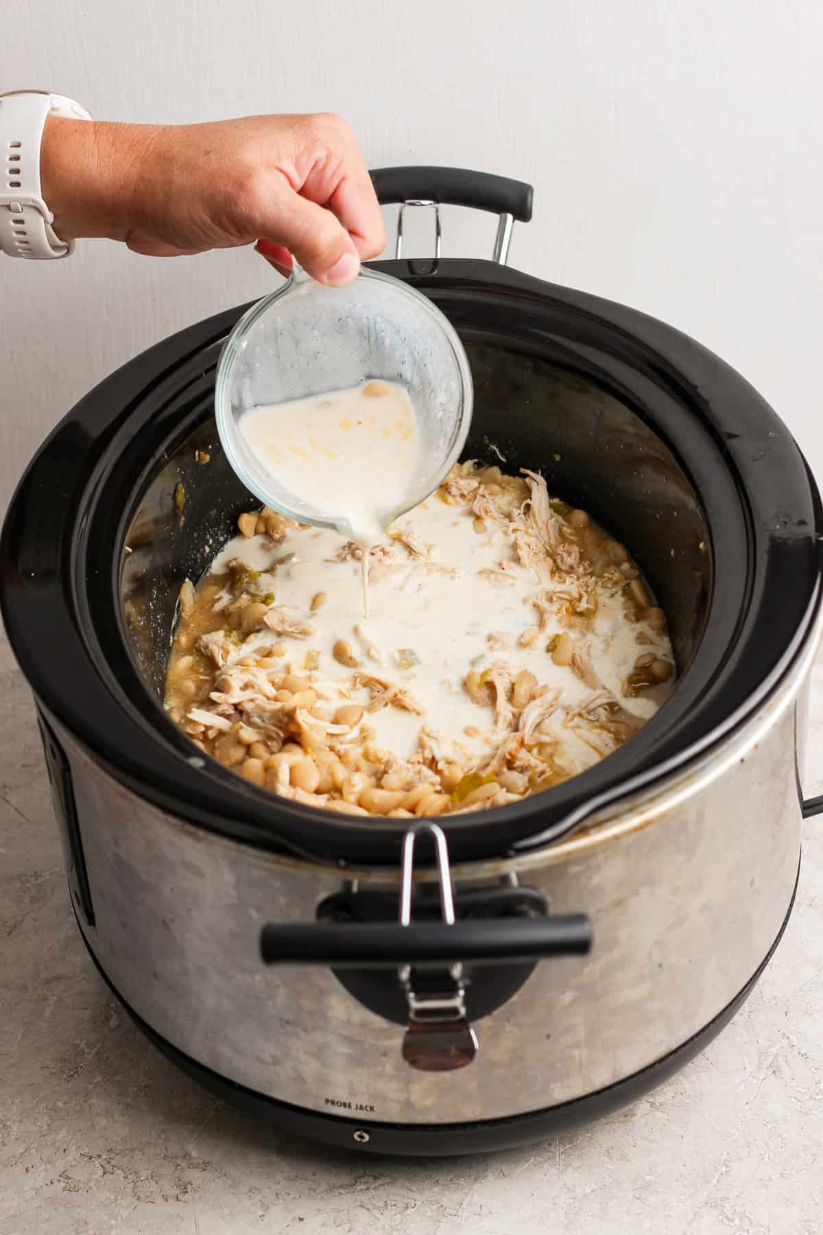 Creamy cottage cheese mixture being poured into a slow cooker of shredded chicken and white beans.