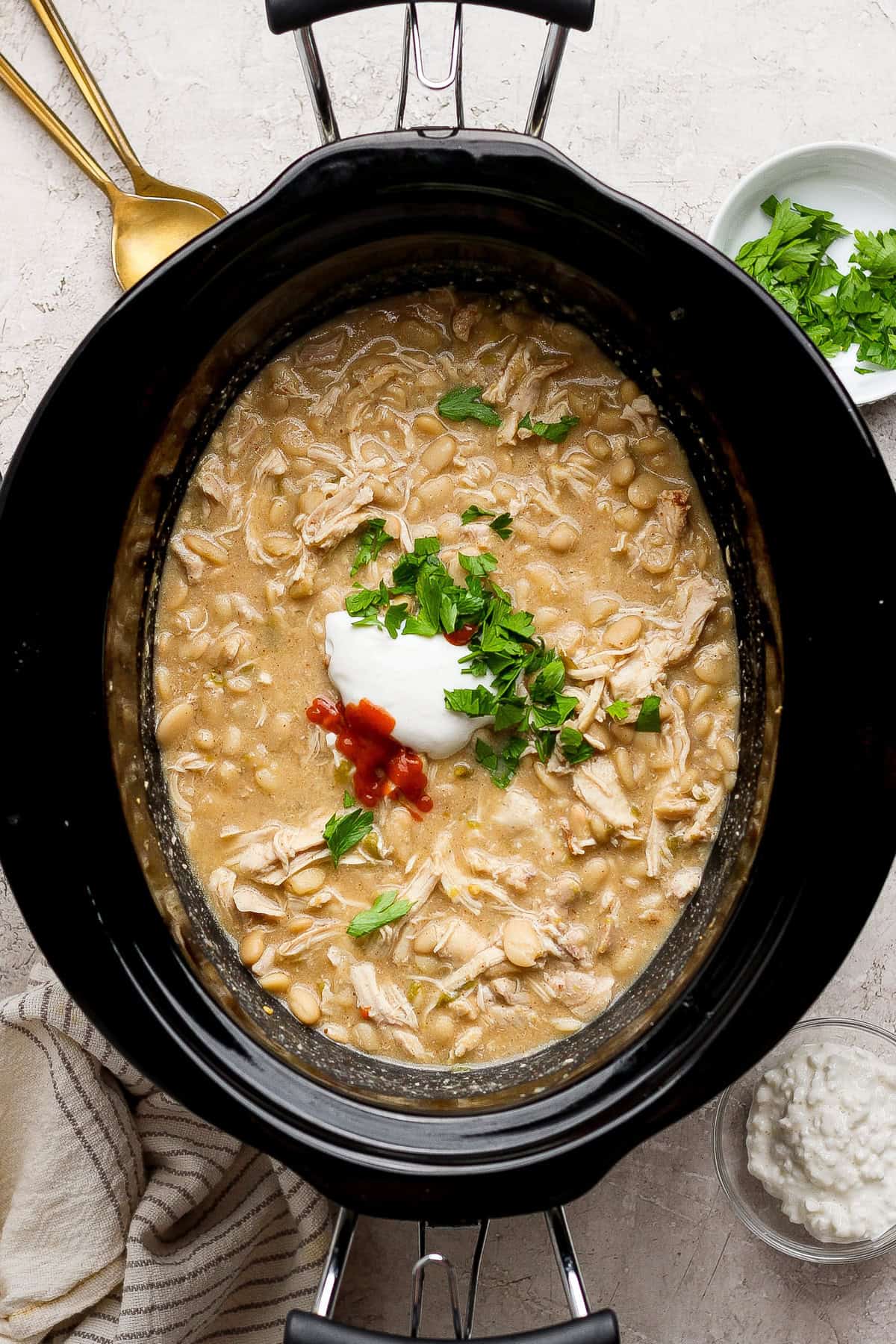 Creamy white chicken chili in a slow cooker topped with cottage cheese, hot sauce, and fresh herbs.