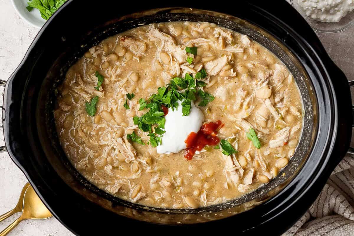 Slow cooker filled with white chicken chili topped with cottage cheese, hot sauce, and chopped fresh herbs.