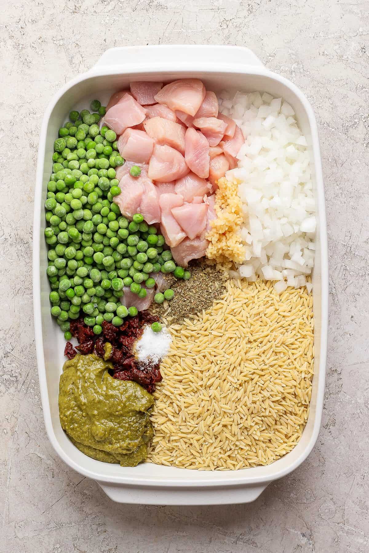 Raw chicken, dry orzo, frozen peas, onion, garlic, pesto, sun-dried tomatoes, and seasonings arranged in a white casserole dish.