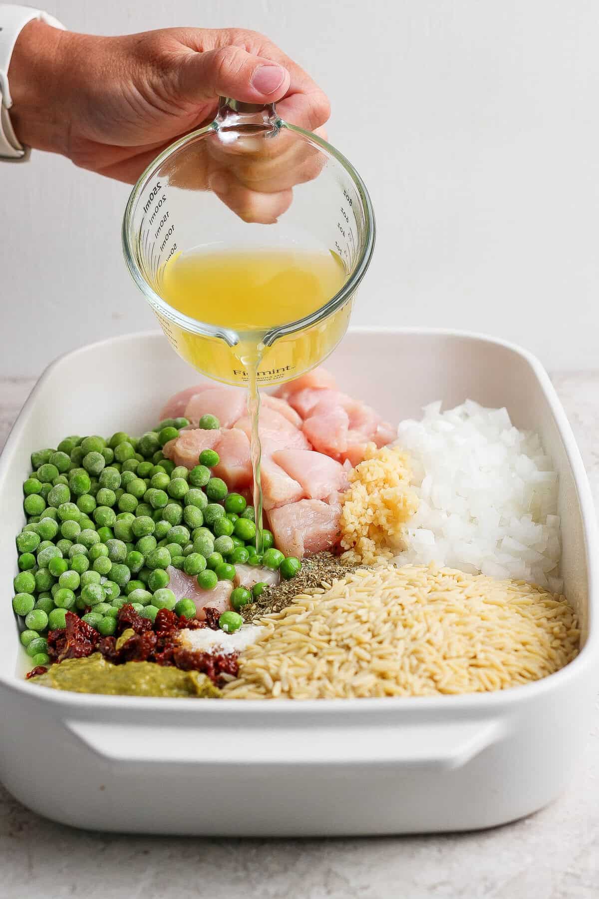Chicken broth being poured over raw chicken, dry orzo, peas, pesto, sun-dried tomatoes, onion, garlic, and seasonings in a casserole dish.