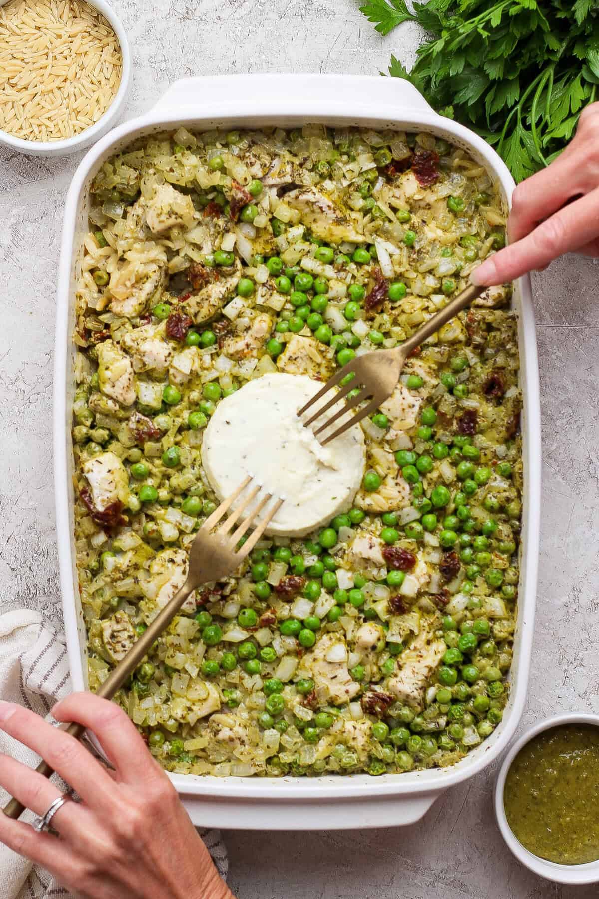 Hands using forks to mix softened Boursin into baked chicken pesto orzo with peas and sun-dried tomatoes.