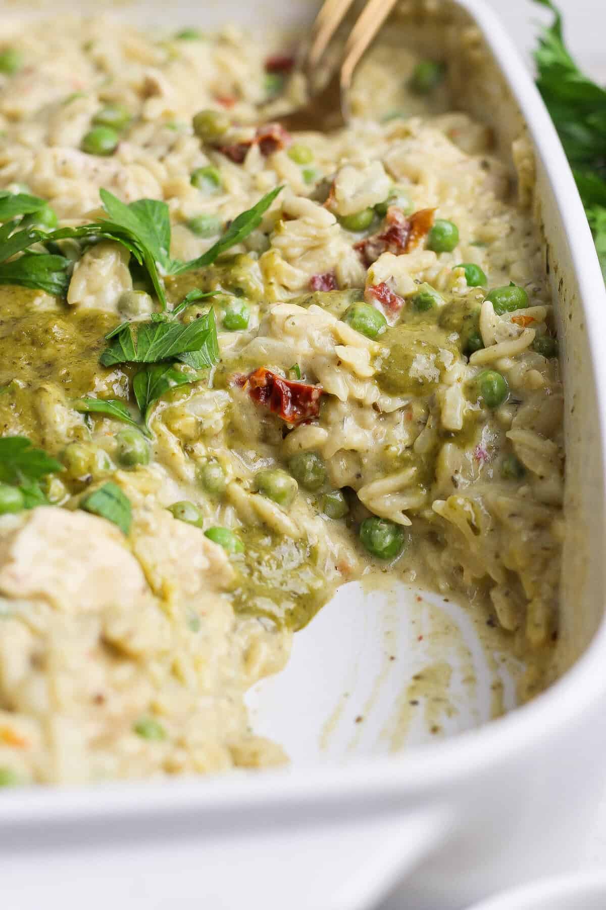 A serving spoon scooping creamy chicken pesto orzo with peas, sun-dried tomatoes, pesto, and fresh parsley from a white casserole dish.