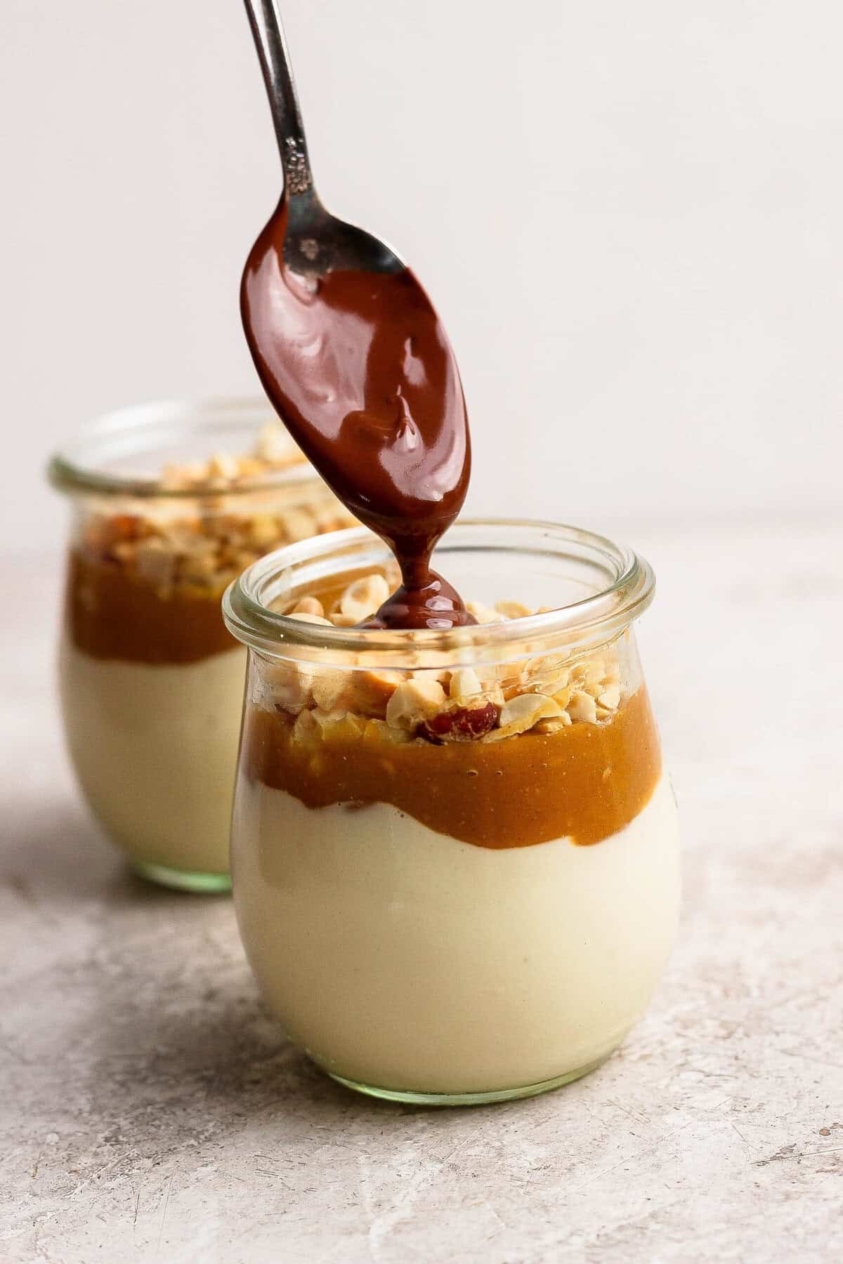 Melted chocolate magic shell being spooned over peanut butter and chopped peanuts in individual cheesecake cups.