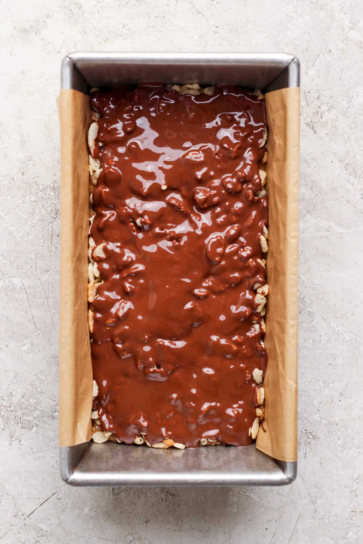 Melted chocolate spread over the crispy rice cereal in a parchment-lined loaf pan.