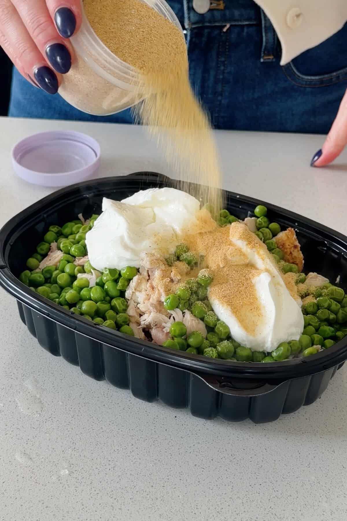 Seasoning sprinkled over chopped chicken, peas, and yogurt in a dark container.