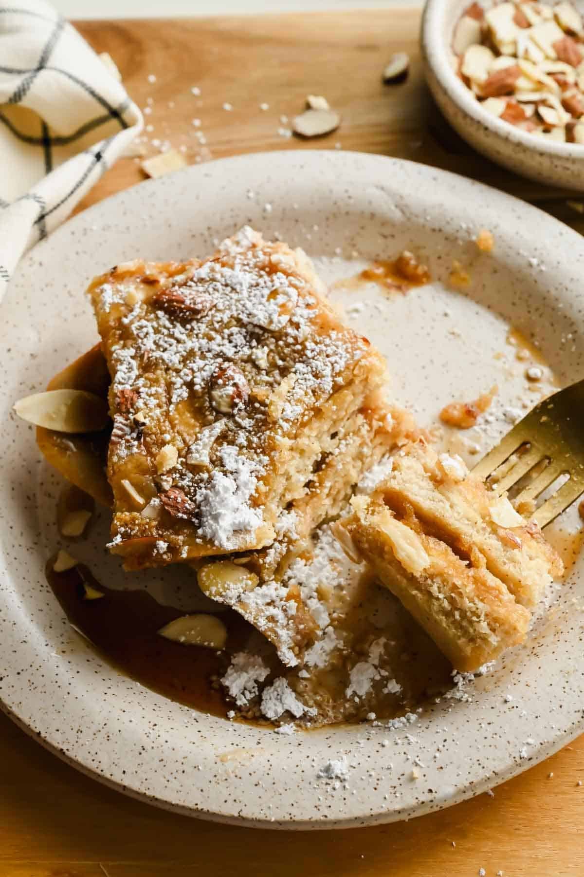 Stacked sheet pan almond croissant pancakes on a plate with maple syrup and powdered sugar.