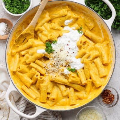Creamy pumpkin rigatoni in a skillet with cottage cheese, Parmesan, red pepper flakes, and green herbs on top.