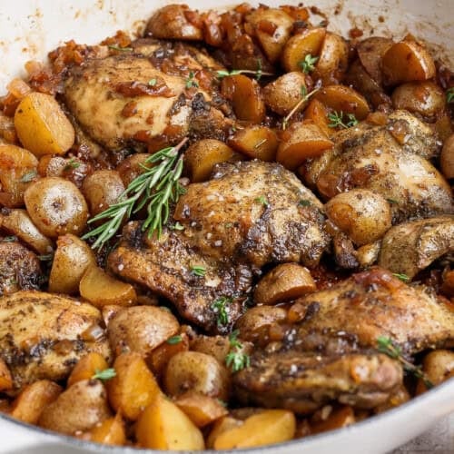 Saucy apple cider chicken thighs with golden baby potatoes, onion, and fresh rosemary in a white skillet.