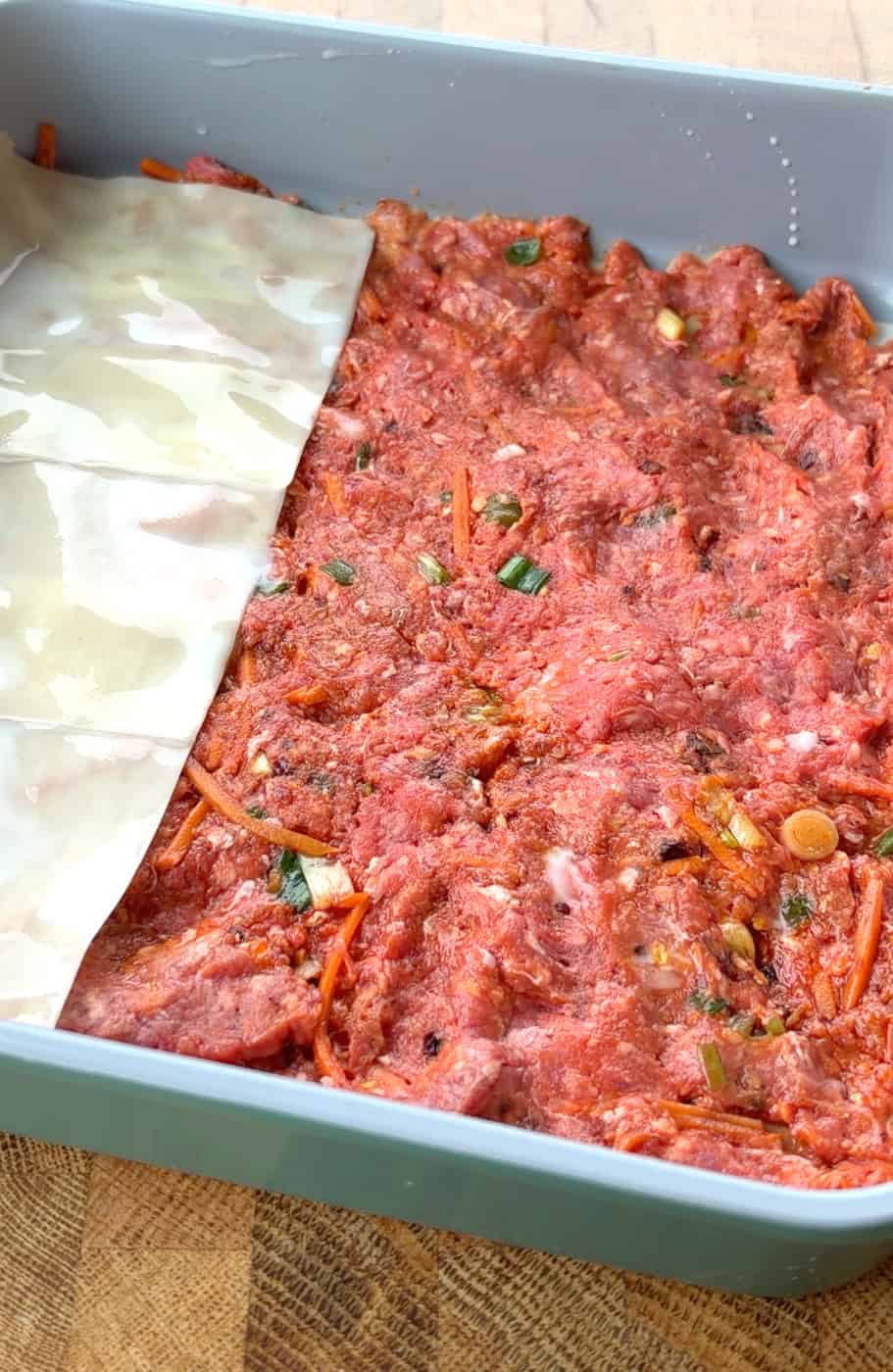 Overlapping wonton wrappers being layered over raw beef filling in a baking pan