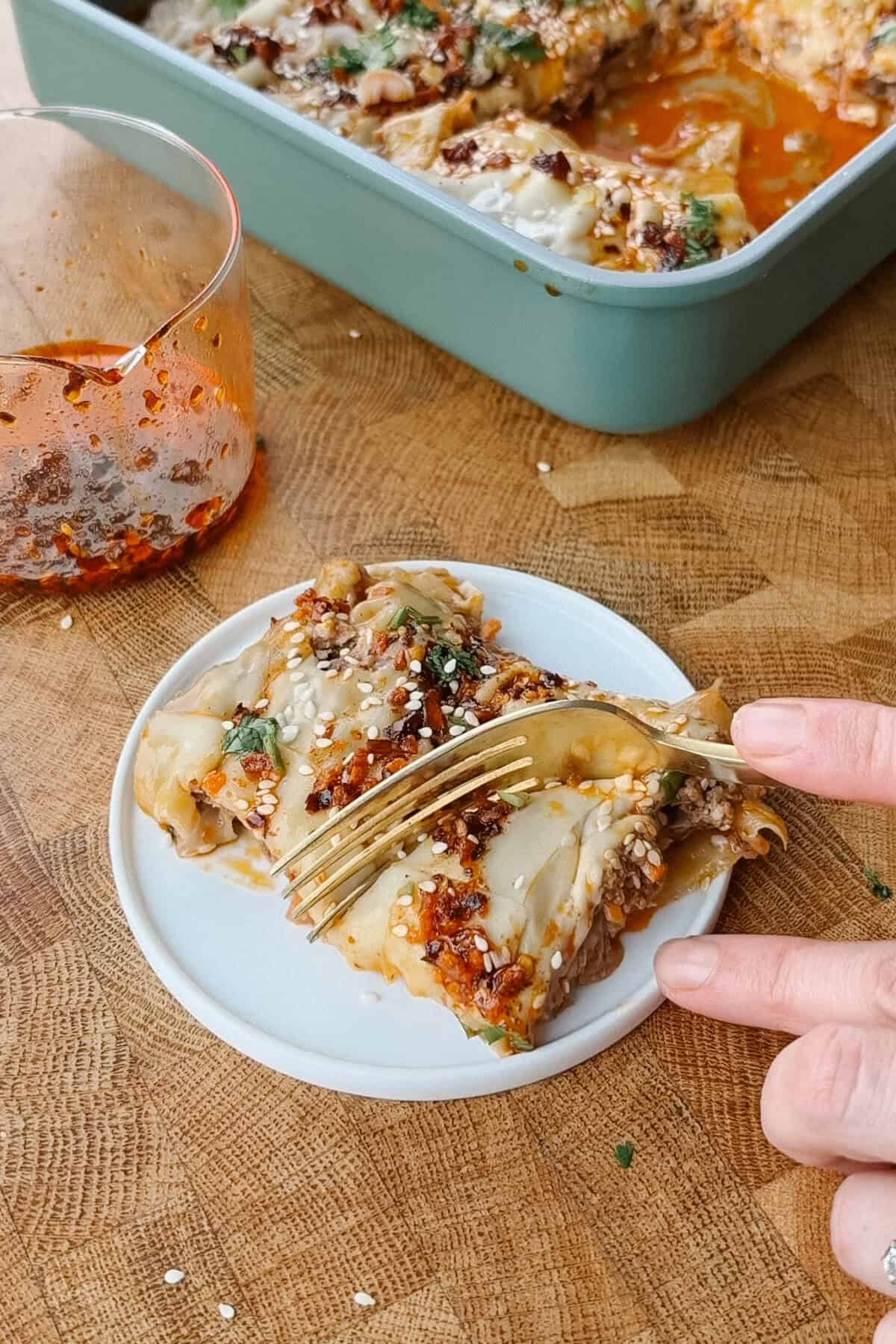 A fork cuts into a serving of dumpling lasagna on a small plate, with a baking dish and a jar of chili oil nearby.