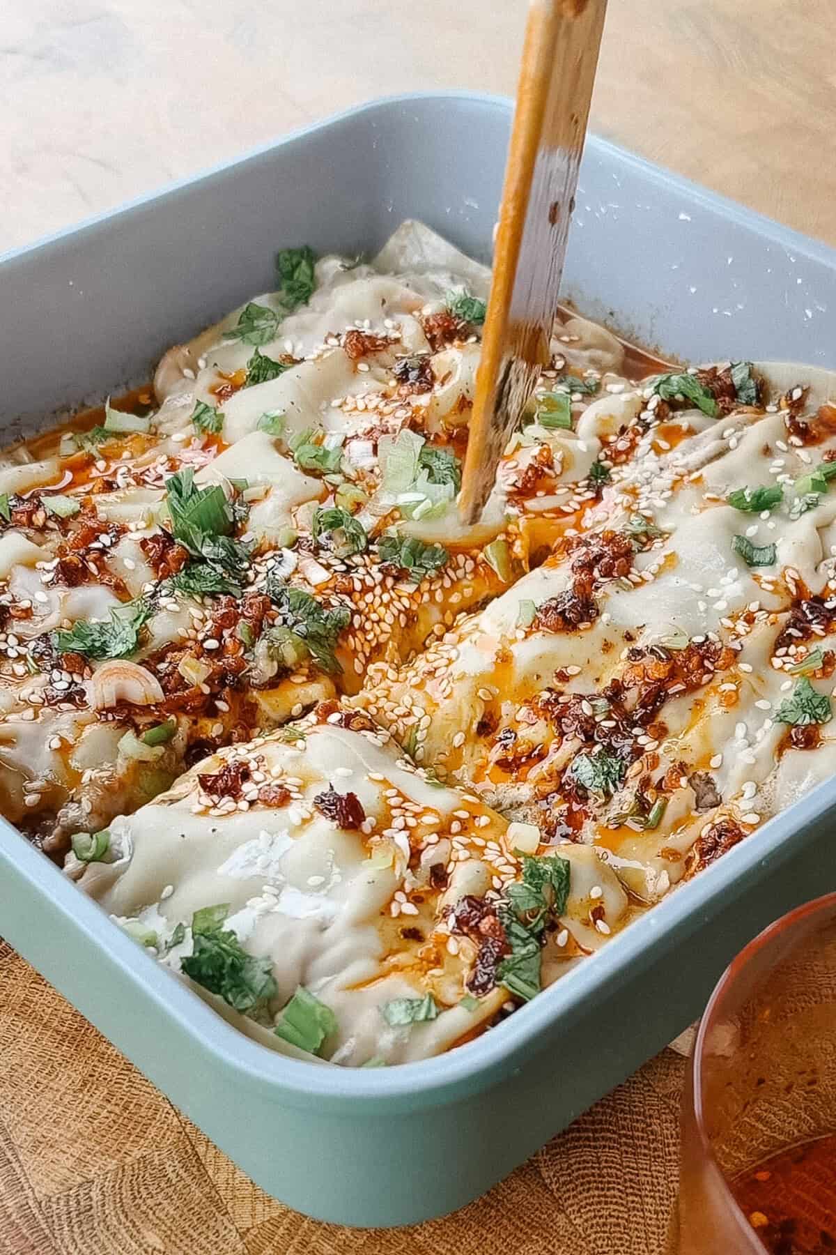 A baking dish of dumpling lasagna topped with chili oil, sesame seeds, and chopped scallions, with a wooden utensil pressing into the dish.