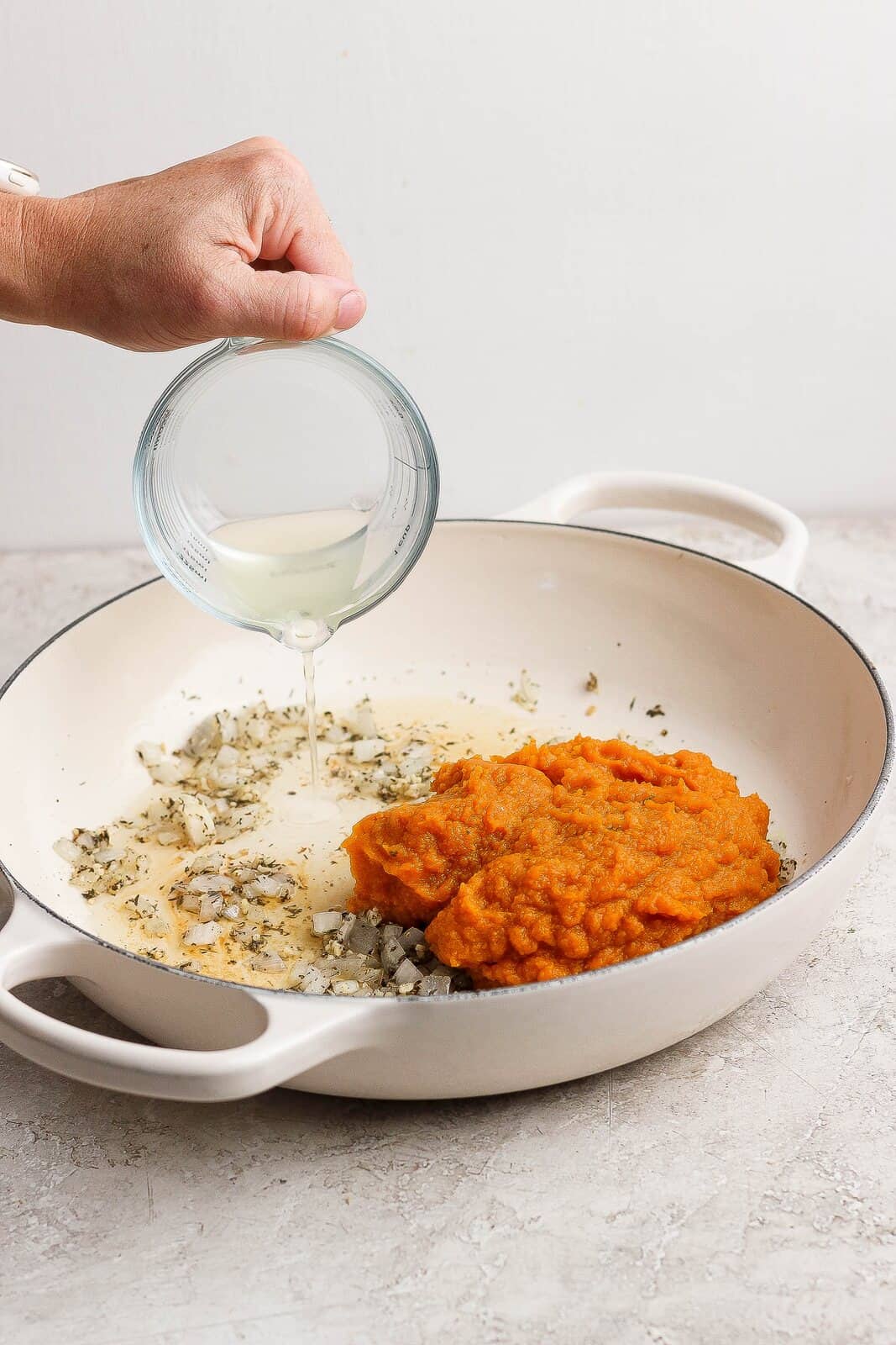 Reserved pasta water being poured into a skillet with pumpkin puree and sauteed aromatics.