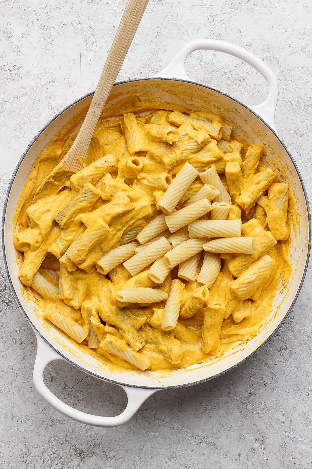 Rigatoni being stirred into creamy pumpkin and cottage cheese sauce in a white skillet.