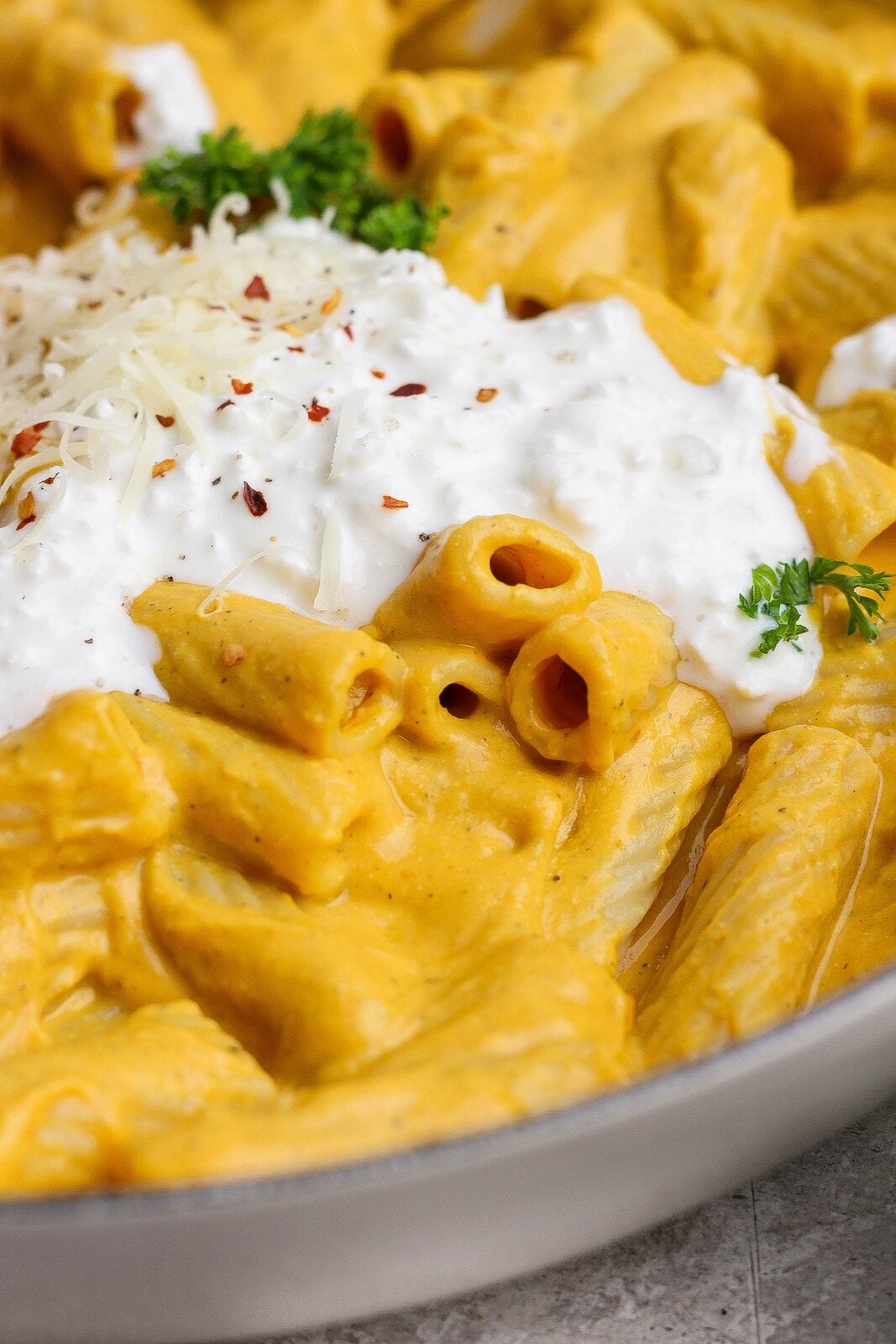 Close-up of creamy pumpkin rigatoni topped with cottage cheese, Parmesan, red pepper flakes, and green herbs.