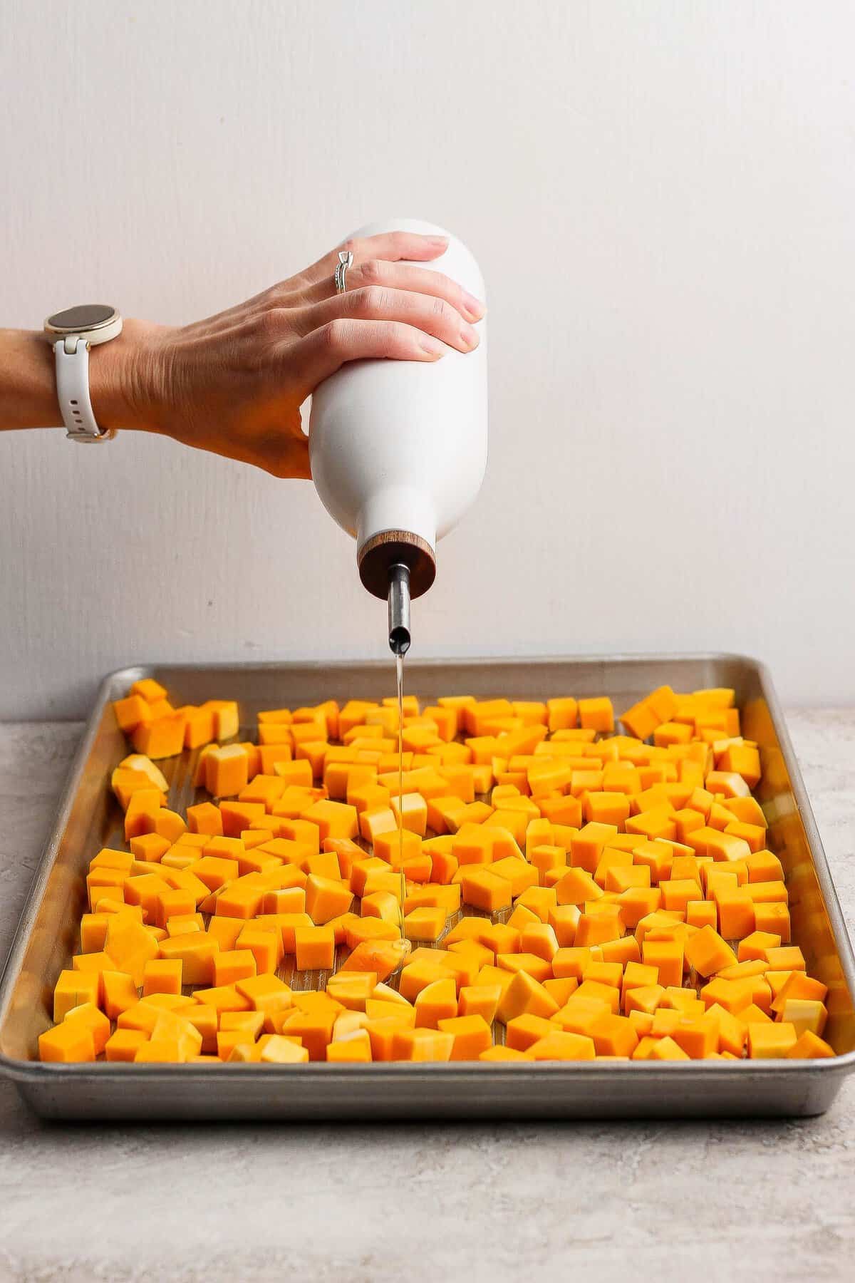Olive oil being poured over cubed butternut squash on a baking sheet before roasting.