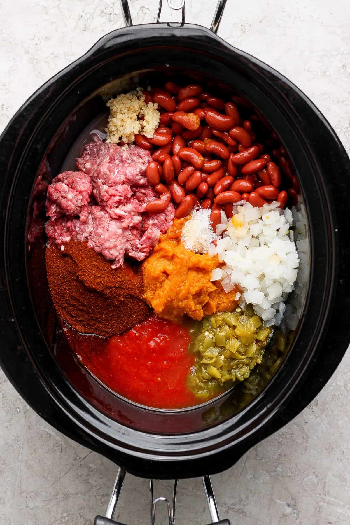Raw ground beef, kidney beans, pumpkin puree, tomatoes, green chiles, onion, garlic, and chili powder arranged in a slow cooker.