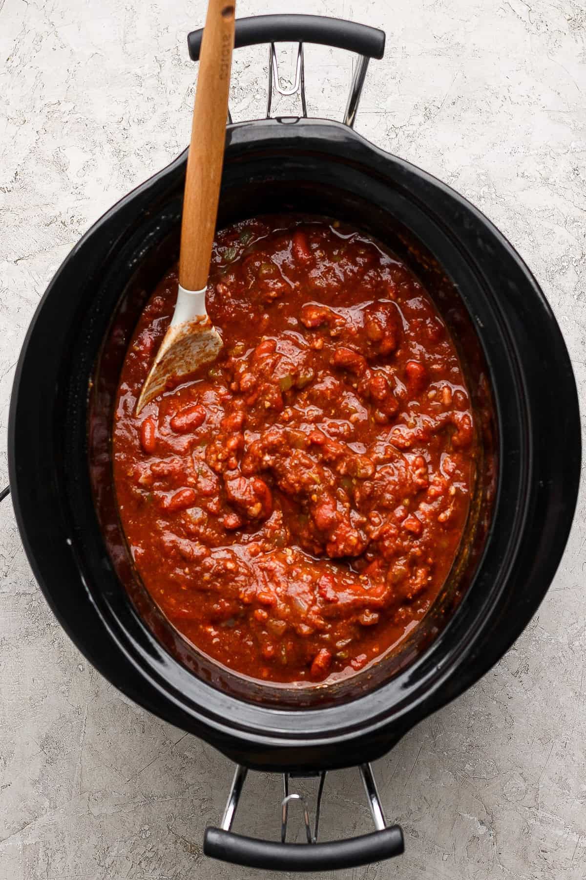 Wooden spoon stirring pumpkin chili with ground beef and kidney beans in a slow cooker.