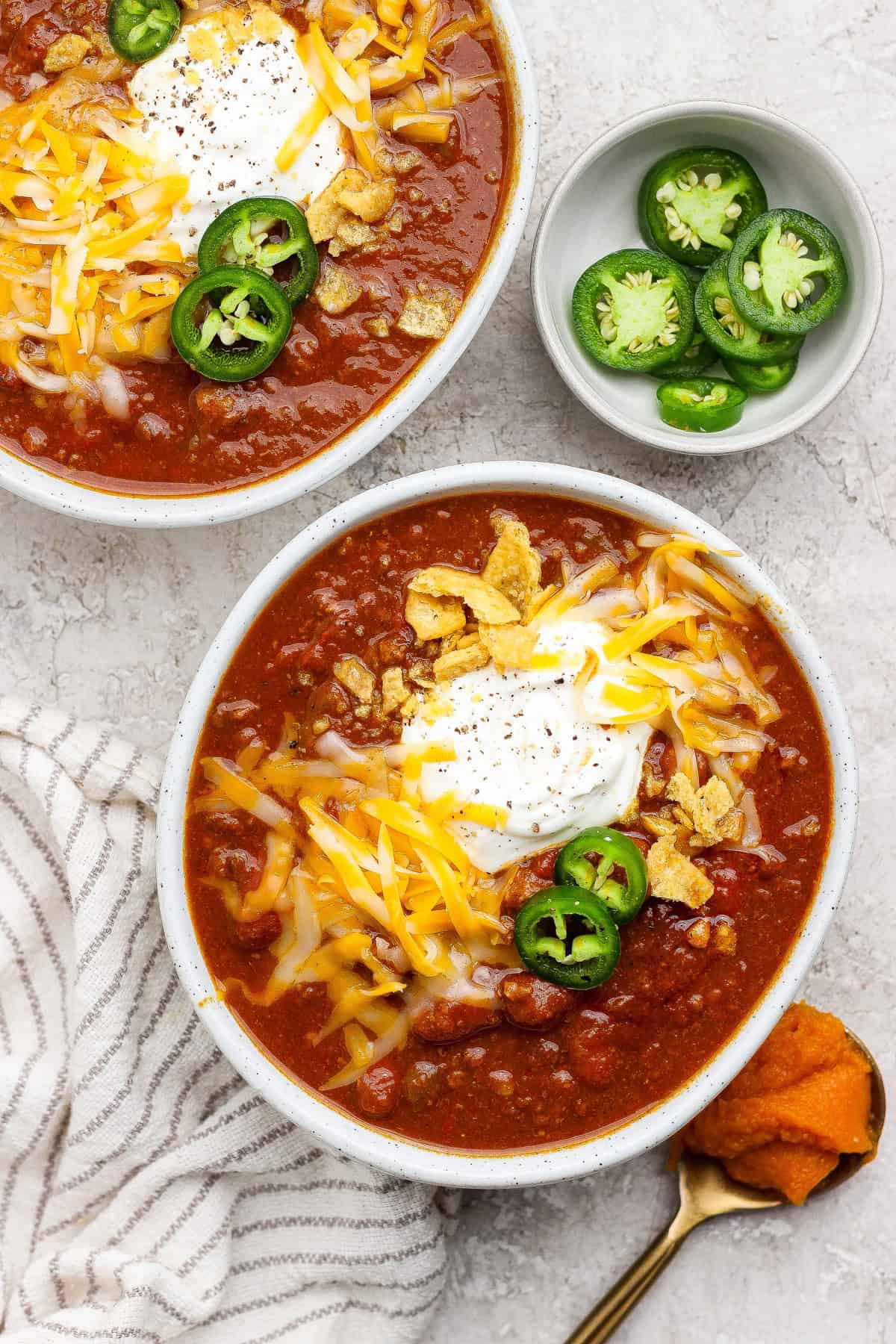 Two bowls of slow cooker pumpkin chili topped with sour cream, shredded cheese, sliced jalape&ntilde;os, and tortilla strips.