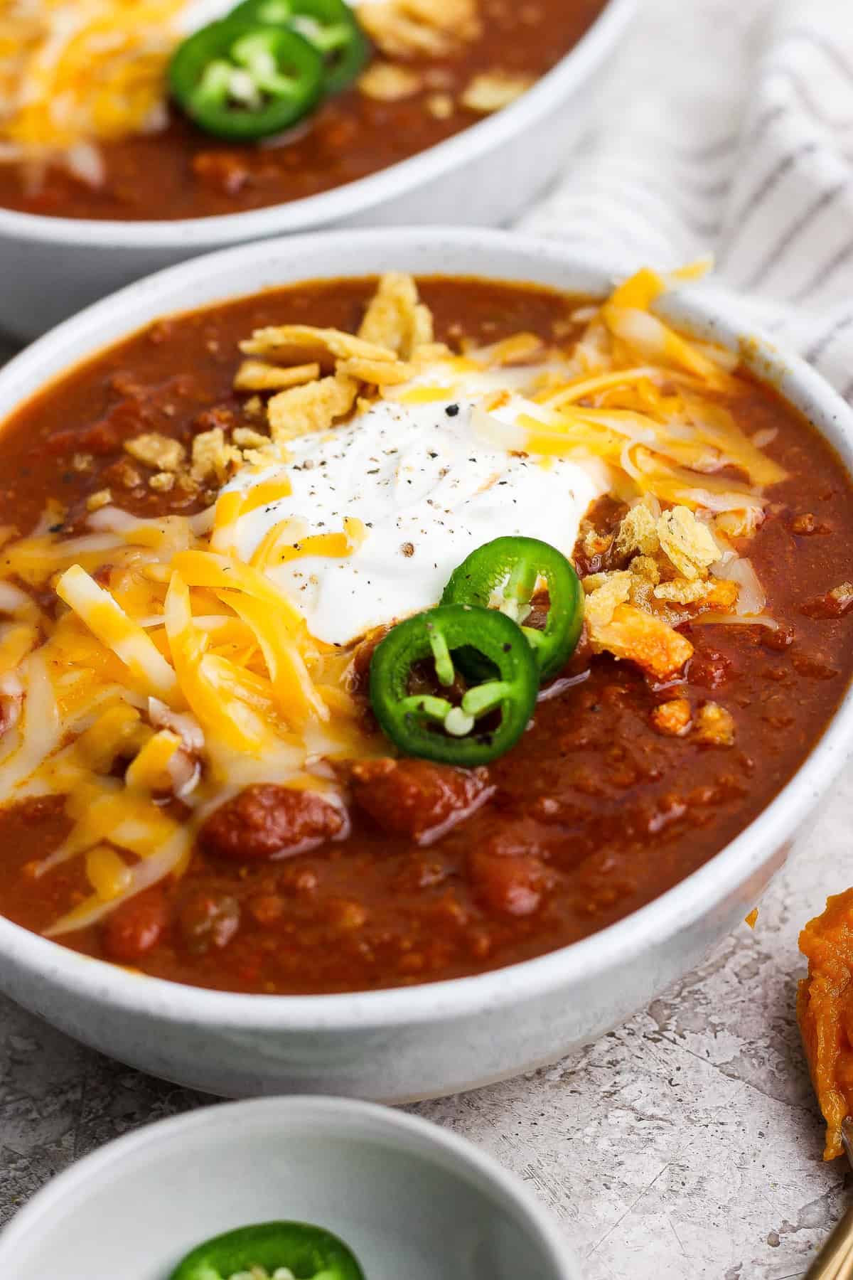 Close-up bowl of slow cooker pumpkin chili with ground beef and kidney beans, topped with sour cream, shredded cheese, jalape&ntilde;o, and tortilla strips.