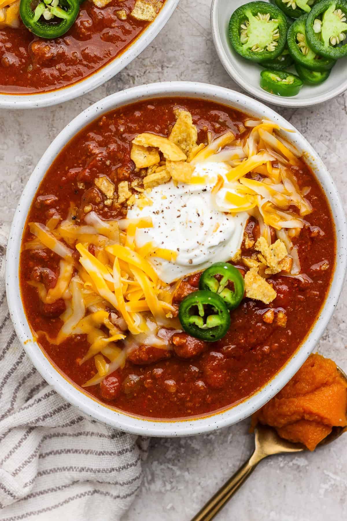 Bowl of pumpkin chili with beef and kidney beans topped with sour cream, shredded cheese, jalape&ntilde;o slices, and tortilla strips.