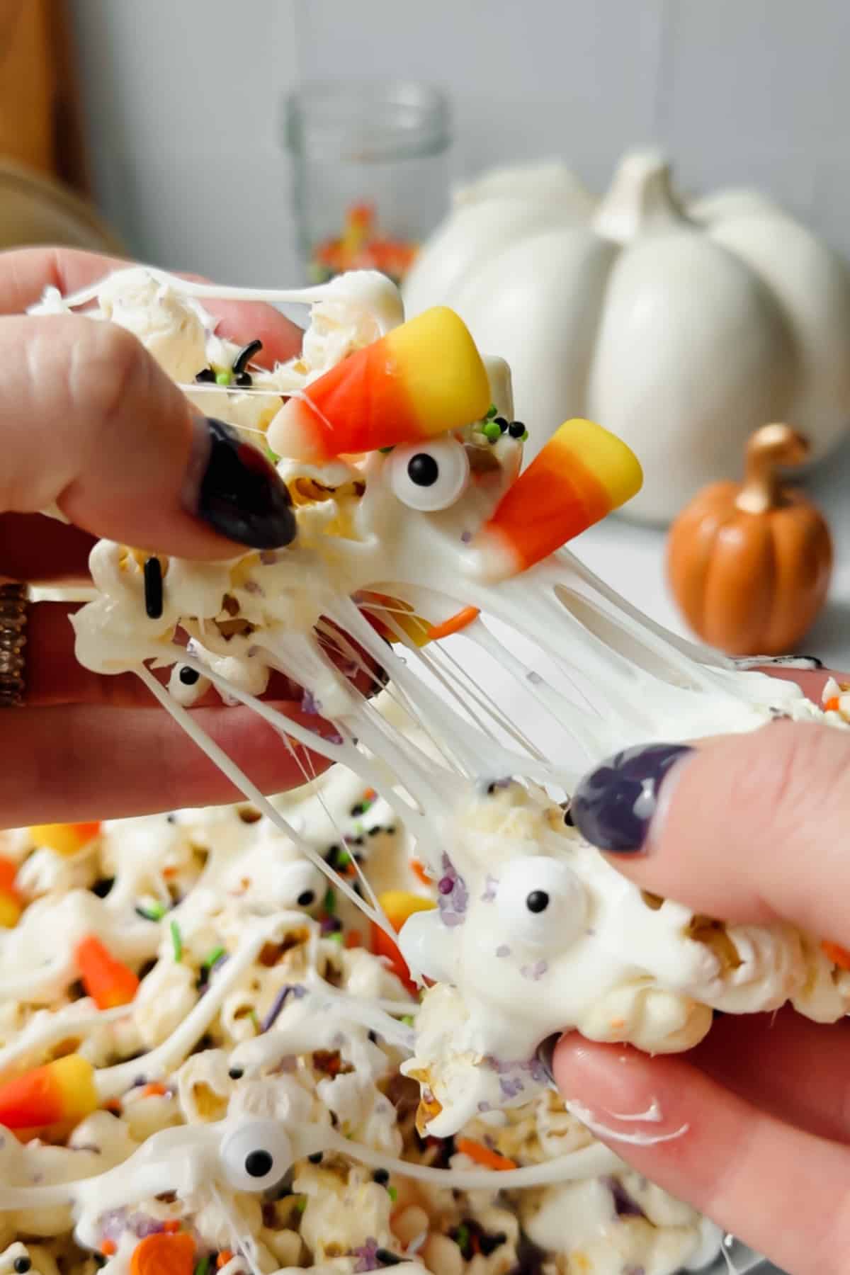 Hands pulling apart marshmallow-coated Halloween popcorn topped with candy corn, sprinkles, and edible candy eyes.