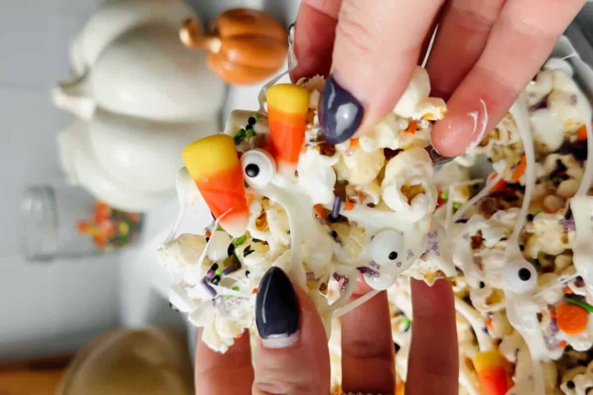 Hands holding a sticky cluster of Halloween popcorn with marshmallow drizzle, candy corn, sprinkles, and edible candy eyes.