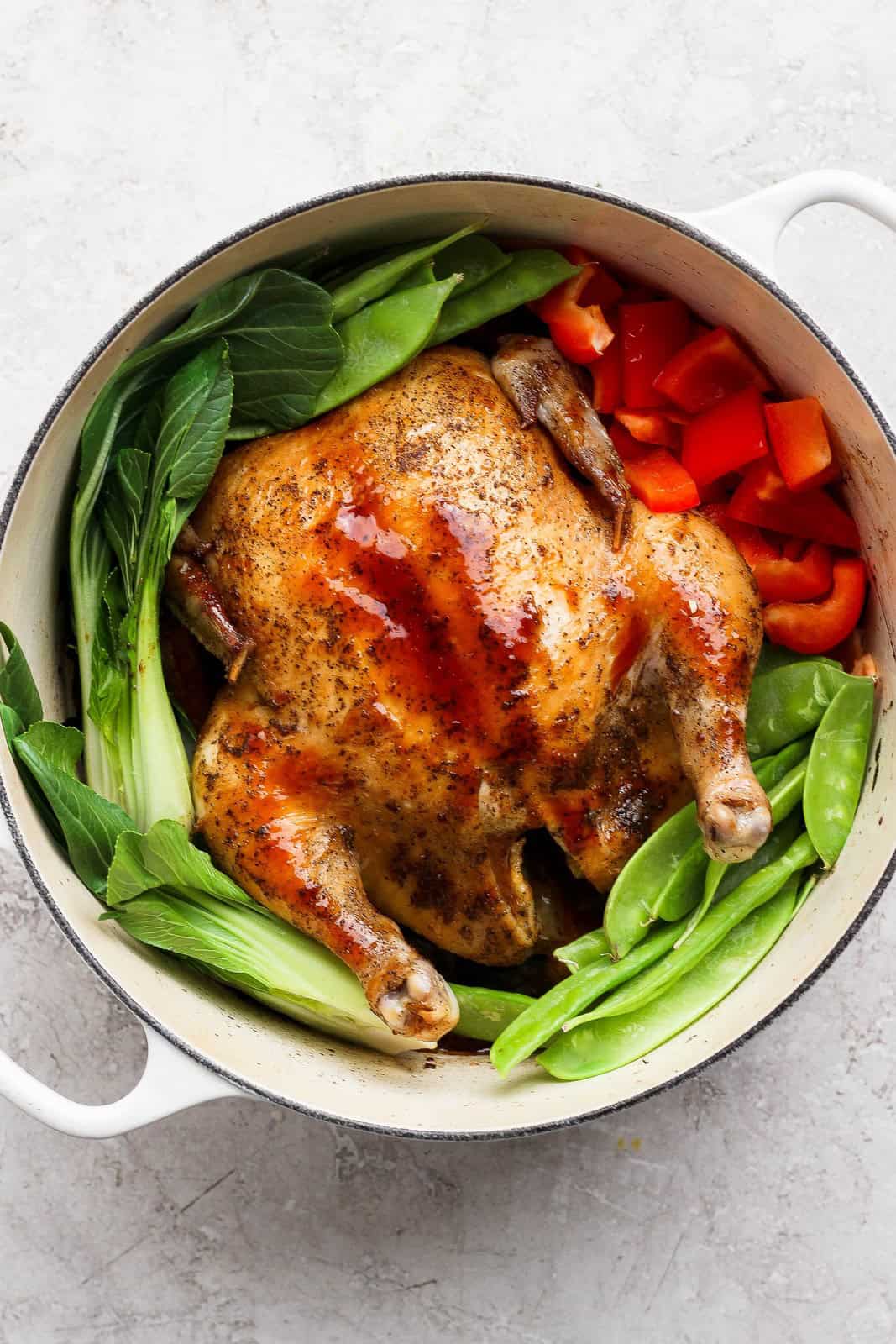 Whole chicken in a Dutch oven surrounded by snow peas, bok choy, and red bell pepper.