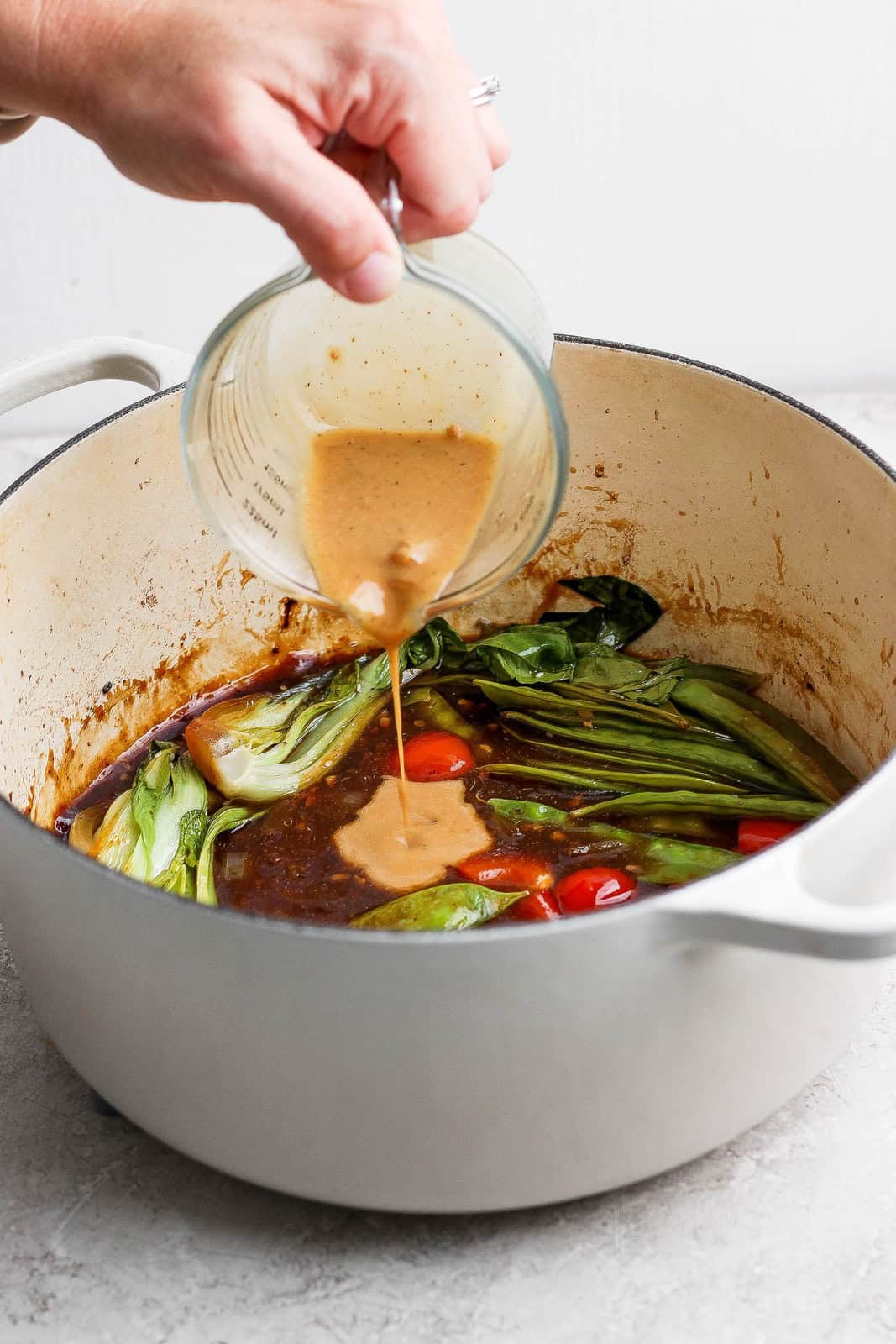 A hand pours a thickened sauce into a Dutch oven of bok choy, red bell pepper, and stir-fry sauce.