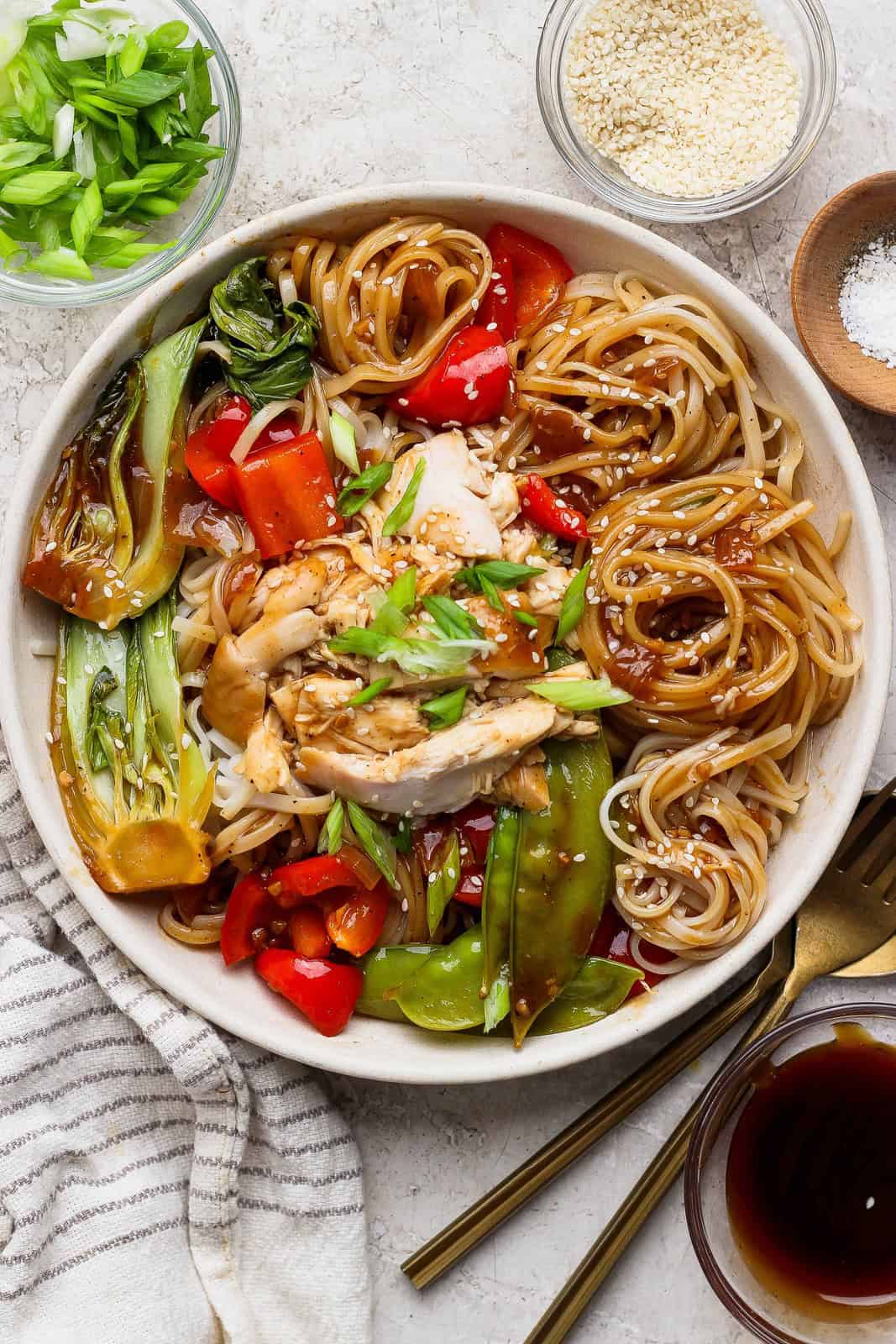 Finished bowl of chicken and vegetable stir-fry with glossy noodles, bok choy, red bell pepper, and sesame seeds.