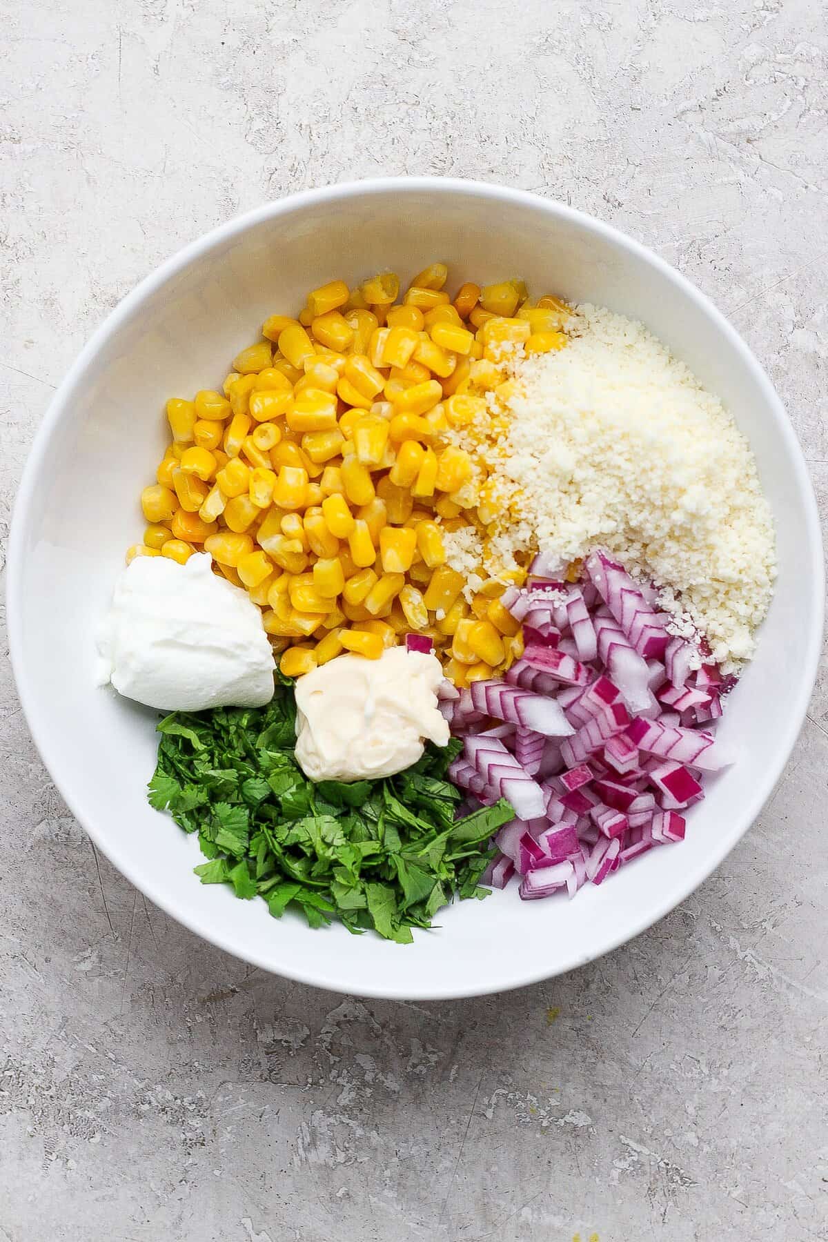 Charred corn, cotija, cilantro, red onion, lime juice, crema, and mayonnaise in a bowl before mixing.