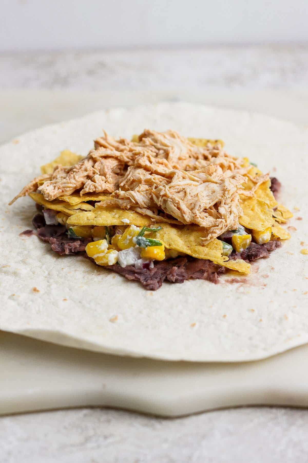 A large flour tortilla layered with smashed black beans, street corn salad, tortilla chips, and creamy shredded chicken.