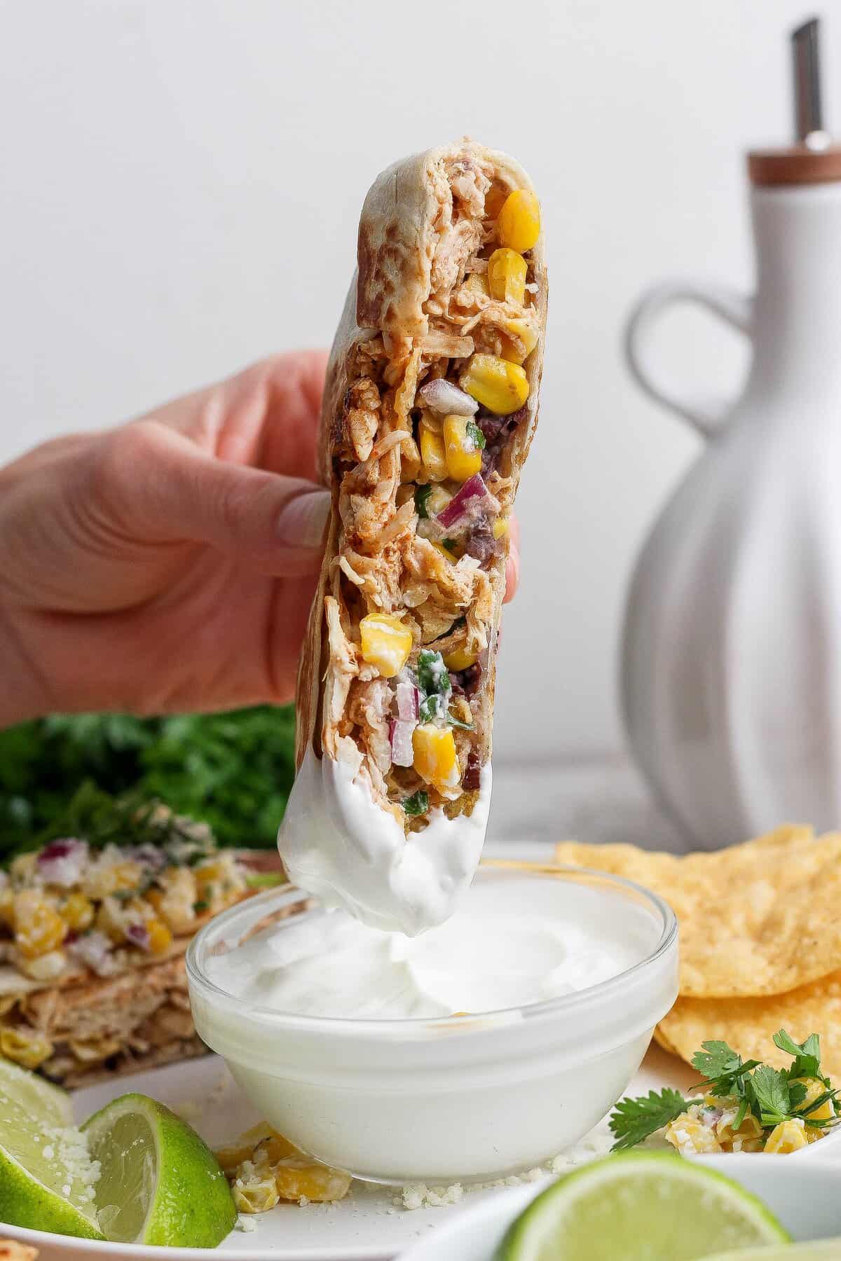 A halved street corn chicken crunchwrap dipped into a bowl of crema.